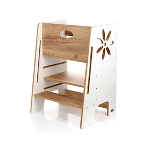 Può includere: Uno sgabello a gradini bianco e color legno a tre livelli. La sezione superiore e i ripiani hanno una finitura a venature del legno, mentre i lati sono bianchi. Un ritaglio a forma di fiore decora un lato. Lo sgabello è progettato per aiutare i bambini a raggiungere superfici più alte.