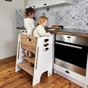 Può includere: Uno sgabello da cucina bianco e color legno con due bambini in piedi sopra. Lo sgabello ha un piano e gradini in legno, con un telaio bianco. I bambini sono vicino a un fornello e un piano di lavoro con cibo.