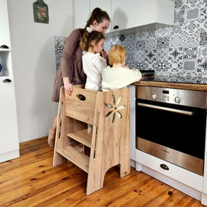 Può includere: Uno sgabello in legno con un ritaglio a forma di fiore sul lato. Lo sgabello ha due gradini e una piattaforma superiore. Il legno ha una venatura naturale. Lo sgabello è in una cucina con una donna e due bambini.