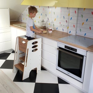 Puede incluir: Un taburete de cocina blanco y con efecto madera con tres recortes ovalados en el lateral. El taburete tiene dos escalones y una plataforma. La cocina tiene armarios blancos, suelo ajedrezado en blanco y negro y un salpicadero con diseños de pájaros de colores.