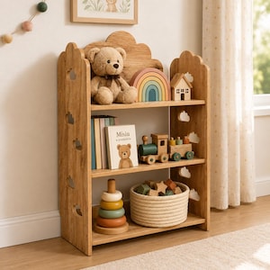 Op de afbeelding: Een lichtbruine houten boekenplank met drie niveaus en wolkvormige uitsparingen aan de zijkanten. De planken bevatten een teddybeer, een regenboogstapelaar, een houten huisje, boeken waaronder een met de titel Misiu, een houten trein, een ringstapelspeeltje en een touwmand.