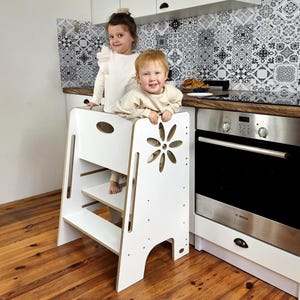 Può includere: Uno sgabello da cucina in legno bianco con un ritaglio a forma di fiore sul lato. Lo sgabello ha due gradini ed è progettato per aiutare i bambini a raggiungere i piani di lavoro. Due bambini sono in piedi sullo sgabello davanti a un piano cucina.