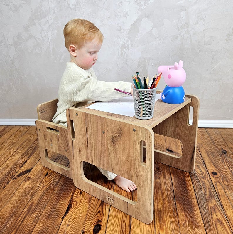 May include: A wooden children's desk and chair set with a light wood grain finish. A young child is seated at the desk, drawing with colored pencils. A pink and blue Peppa Pig figurine sits on the desk. The desk is on a wooden floor.