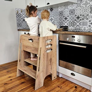 Può includere: Uno sgabello da cucina in legno con finitura a venatura naturale. Lo sgabello ha due gradini e una barra di sicurezza. Il design include ritagli ovali sul lato. Lo sgabello è progettato per aiutare i bambini a raggiungere i piani di lavoro.