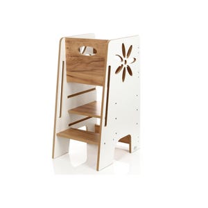 Pode incluir: Uma torre de aprendizagem de madeira com exterior branco e interior com veios de madeira natural. A estrutura possui dois degraus, uma vedação de segurança no topo com um recorte para pega e um recorte decorativo em forma de flor no painel lateral.