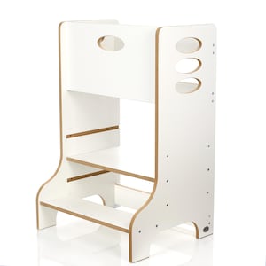 Può includere: Uno sgabello in legno bianco con finiture in legno naturale. Lo sgabello ha due gradini e un'apertura per la maniglia nella parte superiore. Il pannello laterale presenta tre ritagli ovali. Il design è semplice e funzionale, adatto ai bambini.