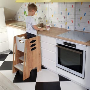 Puede incluir: Un taburete de cocina de madera con un acabado de veta de madera clara. El taburete tiene dos escalones y un panel trasero blanco. Un niño está de pie sobre el taburete en una encimera de cocina. La cocina tiene armarios blancos, suelo a cuadros blanco y negro y un horno blanco.