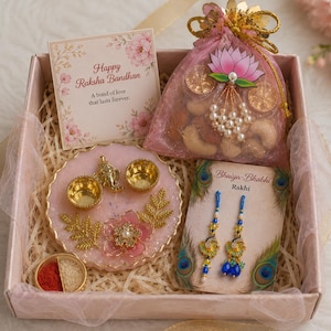 Può includere: Una confezione regalo per Raksha Bandhan contenente un sacchetto in organza rosa con noci, un biglietto con la scritta Happy Raksha Bandhan, un vassoio rotondo in resina rosa con due ciotole dorate e una statuetta di Ganesha e due braccialetti.