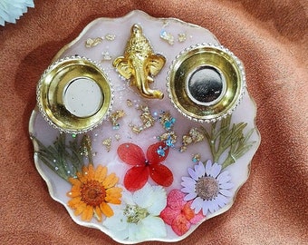May include: A decorative resin tray with a light pink base, gold trim, and embedded dried flowers. It features a gold Ganesh statue and two gold candle holders. The tray is approximately 15 cm in diameter.