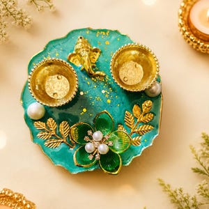 Puede incluir: Una bandeja decorativa de color verde azulado y dorado con un diseño floral. La bandeja presenta dos cuencos dorados, una figura de elefante dorado y una flor verde con detalles de perlas. Acentos de hojas doradas y perlas blancas completan el diseño.