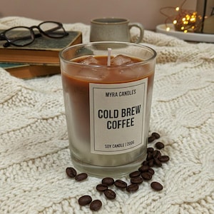Op de afbeelding: Een heldere glazen kaars met een gelaagd ontwerp dat koud gezette koffie imiteert. De kaars heeft een witte basis, een bruin middengedeelte en een bovenkant die op ijsblokjes lijkt. Het etiket luidt "COLD BREW COFFEE" en "MYRA CANDLES". Koffiebonen zijn verspreid.