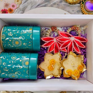 May include: A white gift box contains two teal canisters with floral designs, two red flower-shaped ornaments, and two star-shaped candles. The candles have yellow floral accents. The box is filled with purple shredded paper.