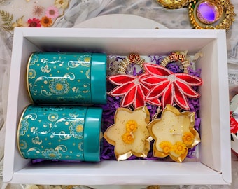 May include: A white gift box contains two teal canisters with floral designs, two red flower-shaped ornaments, and two star-shaped candles. The candles have yellow floral accents. The box is filled with purple shredded paper.