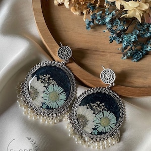 May include: A pair of silver earrings with a round, dark blue resin base. Each earring features a pressed white flower with green accents and a pearl drop fringe.