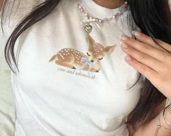 Puede incluir: Camiseta blanca con una ilustración de cervatillo, un lazo azul y el texto "cute and whimsical". El cervatillo es marrón con manchas blancas. También se ve un collar de cuentas con un colgante de corazón.