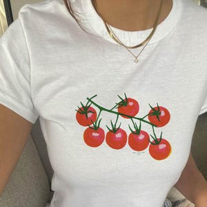 May include: A white short-sleeved t-shirt featuring a graphic print of a cluster of seven bright red tomatoes on a green vine. The tomatoes are arranged in a bunch, and the shirt has a simple crew neckline. The design is centered on the chest.