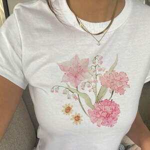 May include: A white short-sleeved t-shirt featuring a watercolor-style floral graphic on the front. The design includes pink lilies, clusters of small white lily of the valley flowers, pink carnations, and two small yellow daisy-like flowers.
