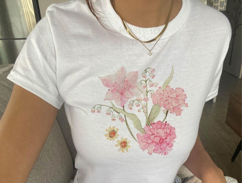 May include: A white short-sleeved t-shirt featuring a watercolor-style floral graphic on the front. The design includes pink lilies, clusters of small white lily of the valley flowers, pink carnations, and two small yellow daisy-like flowers.
