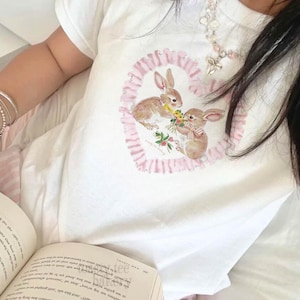 May include: A white short-sleeved t-shirt featuring a central graphic of two brown rabbits exchanging a bouquet of yellow and green flowers. The rabbits are framed by a pink ruffled heart design. The shirt is worn with a beaded necklace.