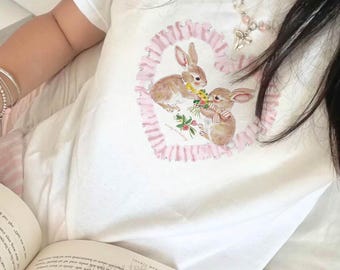 May include: A white short-sleeved t-shirt featuring a central graphic of two brown rabbits exchanging a bouquet of yellow and green flowers. The rabbits are framed by a pink ruffled heart design. The shirt is worn with a beaded necklace.
