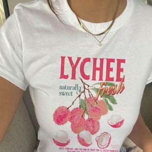 May include: A white short-sleeved t-shirt featuring a graphic print of lychee fruit on a branch. The text reads LYCHEE, naturally sweet, and Fresh. Below the fruit, smaller text reads, Juicy, vibrant, and the kind of fruit you'll catch yourself reaching for again and again.