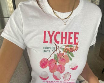 Puede incluir: Una camiseta blanca de manga corta con un estampado gráfico de lichis en una rama. El texto dice LYCHEE, naturally sweet y Fresh. Debajo de la fruta, un texto más pequeño dice: Juicy, vibrant, and the kind of fruit you'll catch yourself reaching for again and again.