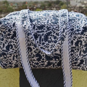 May include: A rectangular quilted duffel bag featuring a dark blue floral and botanical print on a white background. The bag includes two long, sturdy fabric handles with a matching blue and white striped edge detail and a quilted texture throughout.