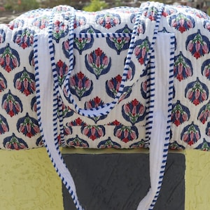 May include: A quilted tote bag with a floral pattern in red, blue, and pink on a cream background. The bag has white handles with blue stripes and a matching trim. The bag is designed for carrying items.