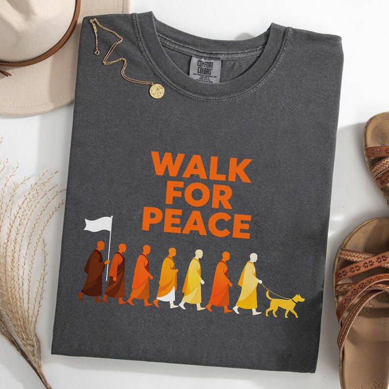 Op de afbeelding: Donkergrijs T-shirt met de oranje tekst "WALK FOR PEACE". Het shirt heeft een afbeelding van mensen in gewaden die met een hond lopen. Een gouden ketting en sandalen zijn zichtbaar.