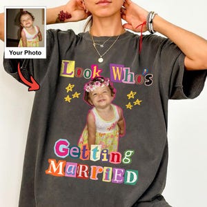 May include: A charcoal gray t-shirt featuring a custom photo print of a young child wearing a floral dress and a flower crown. The text reads Look Who's Getting MARRIED in colorful, blocky, cutout-style letters, accented by yellow star graphics.