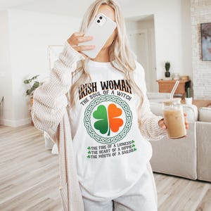 以下が含まれることがあります： 「IRISH WOMAN THE SOUL OF A WITCH」の文字とクローバーのデザインが施された白いTシャツ。クローバーは緑とオレンジで、ケルトノットのデザインで囲まれています。「THE FIRE OF A LIONESS, THE HEART OF A HIPPIE, THE MOUTH OF A SAILOR」の追加テキストが含まれています。