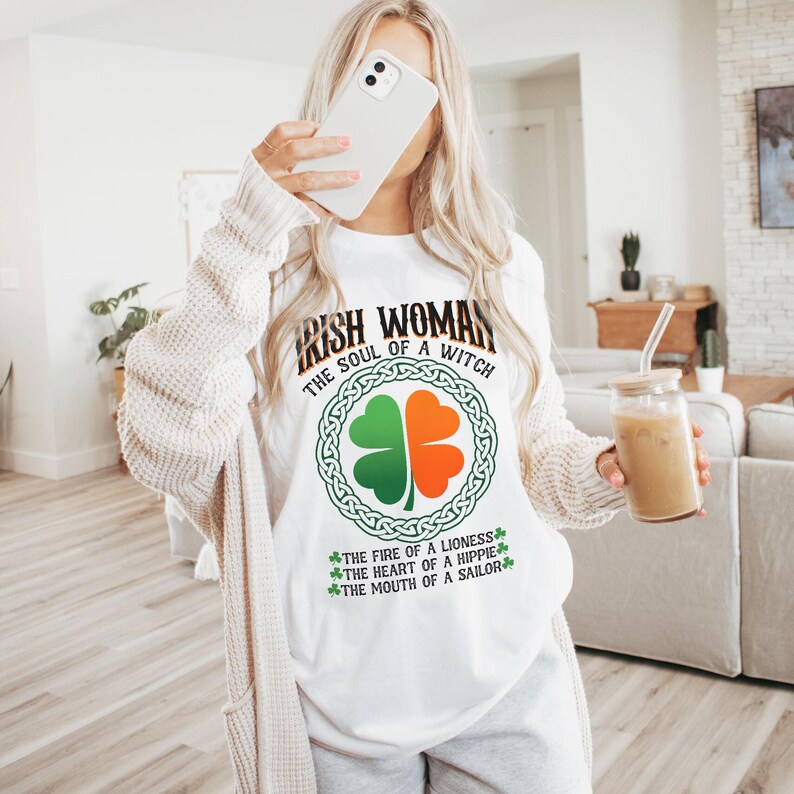 以下が含まれることがあります： 「IRISH WOMAN THE SOUL OF A WITCH」の文字とクローバーのデザインが施された白いTシャツ。クローバーは緑とオレンジで、ケルトノットのデザインで囲まれています。「THE FIRE OF A LIONESS, THE HEART OF A HIPPIE, THE MOUTH OF A SAILOR」の追加テキストが含まれています。