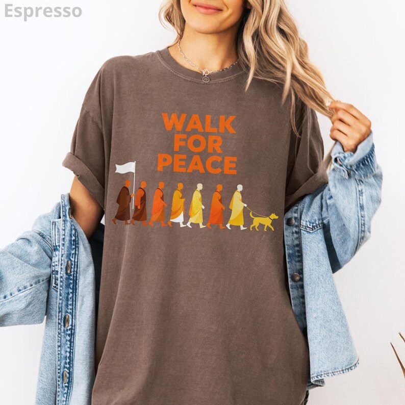 Op de afbeelding: Espresso-kleurig t-shirt met de tekst "WALK FOR PEACE" in oranje. De afbeelding toont een rij figuren in verschillende kleuren, die een witte vlag vasthouden en een hond uitlaten. Het shirt heeft korte mouwen en een ronde hals.
