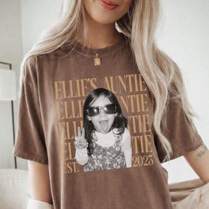 Peut inclure: Un t-shirt marron &agrave; manches courtes avec un motif textuel dor&eacute; r&eacute;p&eacute;t&eacute; indiquant Ellie's Auntie. Au centre, une photographie en noir et blanc d'un enfant portant des lunettes de soleil, faisant un signe de paix et tirant la langue, avec Est. 2023.