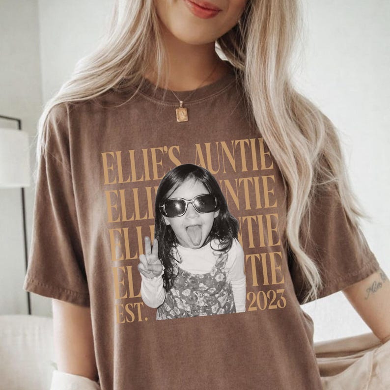 Peut inclure: Un t-shirt marron &agrave; manches courtes avec un motif textuel dor&eacute; r&eacute;p&eacute;t&eacute; indiquant Ellie's Auntie. Au centre, une photographie en noir et blanc d'un enfant portant des lunettes de soleil, faisant un signe de paix et tirant la langue, avec Est. 2023.