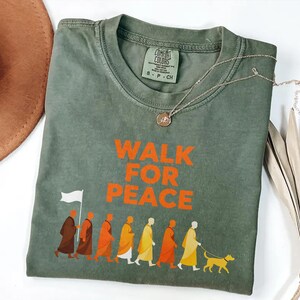 Op de afbeelding: Olijfgroen t-shirt met de oranje tekst "WALK FOR PEACE". Het shirt heeft een afbeelding van mensen in gewaden die met een hond lopen. Een zilveren ketting met een hanger ligt op het shirt.