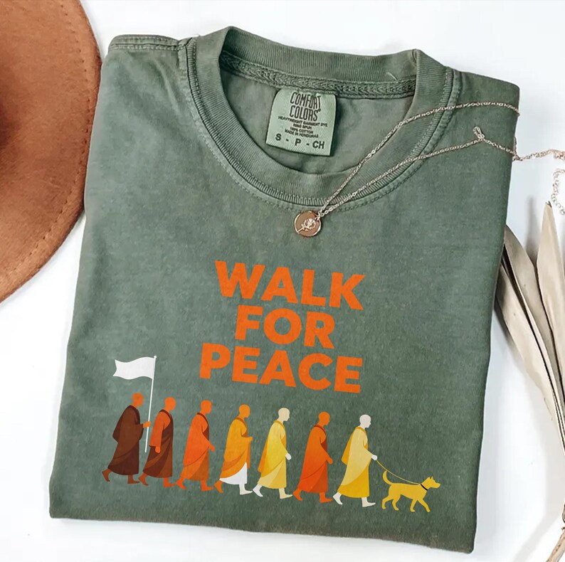 Op de afbeelding: Olijfgroen t-shirt met de oranje tekst "WALK FOR PEACE". Het shirt heeft een afbeelding van mensen in gewaden die met een hond lopen. Een zilveren ketting met een hanger ligt op het shirt.
