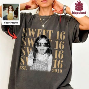 Può includere: Una t-shirt a maniche corte grigio antracite con una grande stampa grafica di una persona che indossa occhiali da sole e tira fuori la lingua. Il testo SWEET 16 è ripetuto verticalmente in lettere dorate, con l'anno 2010 stampato in basso.