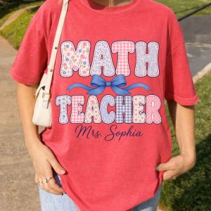 May include: A red short-sleeved t-shirt featuring the text MATH TEACHER in large, patterned block letters with a blue bow graphic in the centre. Below the text, the name Mrs. Sophia is printed in a blue script font. A white bag strap is visible.