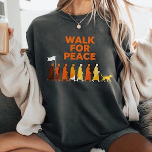 Op de afbeelding: Donkergrijze sweatshirt met de oranje tekst "WALK FOR PEACE". Het ontwerp toont een afbeelding van figuren in gewaden, een hond aan de lijn en een persoon met een witte vlag.
