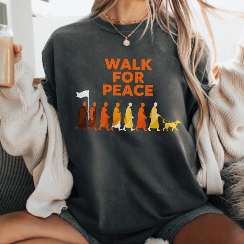 Op de afbeelding: Donkergrijze sweatshirt met de oranje tekst "WALK FOR PEACE". Het ontwerp toont een afbeelding van figuren in gewaden, een hond aan de lijn en een persoon met een witte vlag.