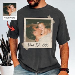 May include: Dark gray t-shirt featuring a vintage-style photo of a parent and child, with the text "Dad Est. 1995." The photo is taped onto the shirt. A smaller photo with the text "Your Photo" is in the upper left corner.