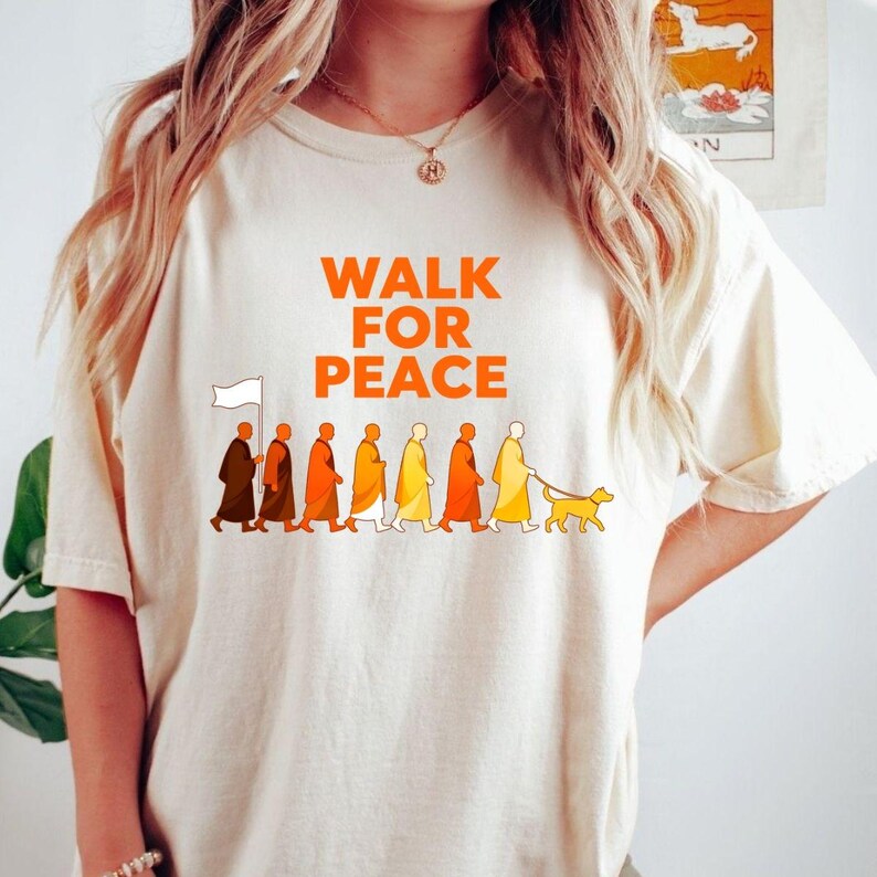 Op de afbeelding: Cr&egrave;mekleurig T-shirt met de oranje tekst "WALK FOR PEACE". Daaronder een afbeelding van mensen in gewaden die met een hond en een witte vlag lopen.