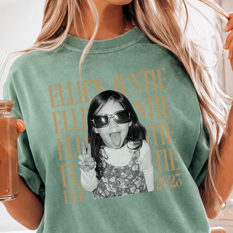 Peut inclure: Un t-shirt &agrave; col rond vert sauge avec une photographie en noir et blanc d'un enfant portant des lunettes de soleil, tirant la langue et faisant un signe de paix. Le texte "ELLIE'S AUNTIE" est r&eacute;p&eacute;t&eacute; en dor&eacute;, avec "EST. 2023" en bas.