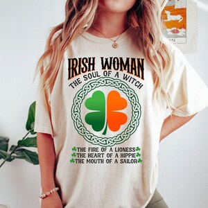 以下が含まれることがあります： 「IRISH WOMAN THE SOUL OF A WITCH」と書かれたクリーム色のTシャツ。緑とオレンジの四つ葉のクローバーのデザインがケルトノットの縁取りの中にあります。「THE FIRE OF A LIONESS」、「THE HEART OF A HIPPIE」、そして「THE MOUTH OF A SAILOR」という言葉も含まれています。