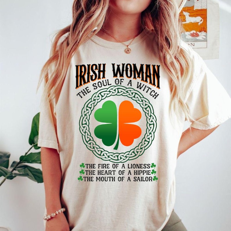 以下が含まれることがあります： 「IRISH WOMAN THE SOUL OF A WITCH」と書かれたクリーム色のTシャツ。緑とオレンジの四つ葉のクローバーのデザインがケルトノットの縁取りの中にあります。「THE FIRE OF A LIONESS」、「THE HEART OF A HIPPIE」、そして「THE MOUTH OF A SAILOR」という言葉も含まれています。