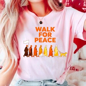 Op de afbeelding: Roze T-shirt met de tekst "WALK FOR PEACE" in oranje. De afbeelding toont een rij figuren in gewaden, die een vlag vasthouden en een hond uitlaten. De figuren zijn in tinten bruin, oranje en geel.