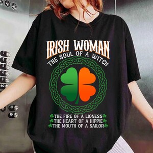 以下が含まれることがあります： 「IRISH WOMAN THE SOUL OF A WITCH」の文字が入った黒いTシャツ。緑とオレンジの四つ葉のクローバーのデザインが中央にあり、緑のケルトノットのボーダーで囲まれています。下部には「THE FIRE OF A LIONESS, THE HEART OF A HIPPIE, THE MOUTH OF A SAILOR」と書かれています。