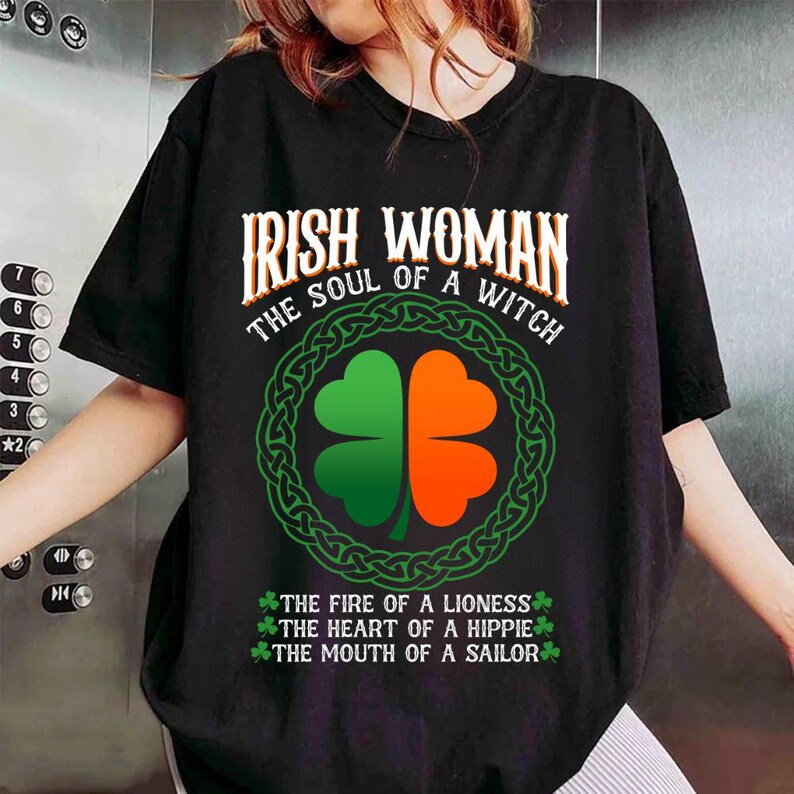 以下が含まれることがあります： 「IRISH WOMAN THE SOUL OF A WITCH」の文字が入った黒いTシャツ。緑とオレンジの四つ葉のクローバーのデザインが中央にあり、緑のケルトノットのボーダーで囲まれています。下部には「THE FIRE OF A LIONESS, THE HEART OF A HIPPIE, THE MOUTH OF A SAILOR」と書かれています。