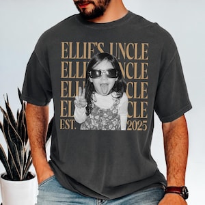 May include: A charcoal gray short-sleeve t-shirt featuring a black and white photograph of a child wearing sunglasses and sticking out their tongue while making a peace sign. The text "ELLIE'S UNCLE" is repeated in tan, with "EST. 2025" at the bottom.
